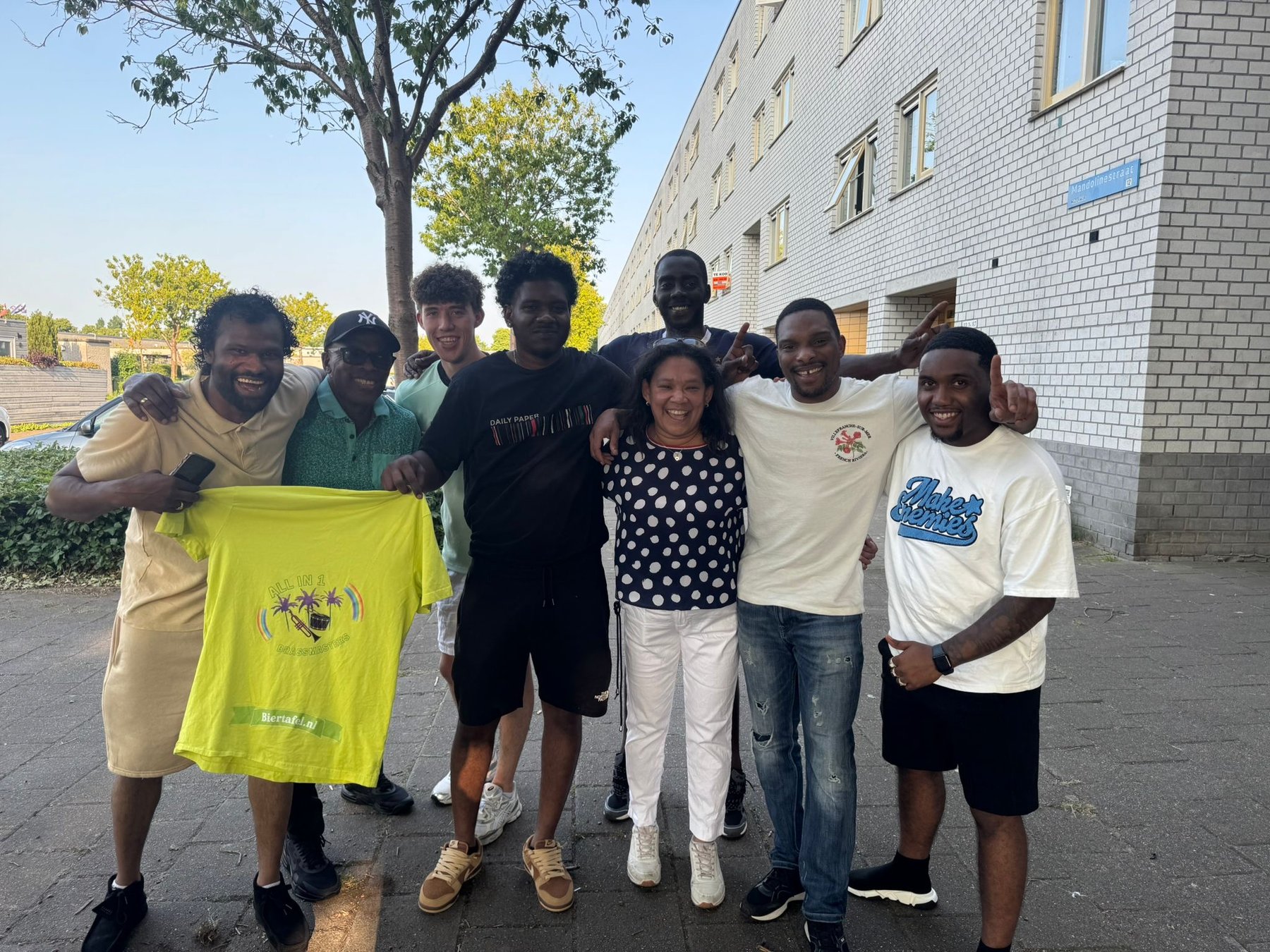 Het team van All In 1 Music Brassband samen op straat, met hun eigen bandshirt