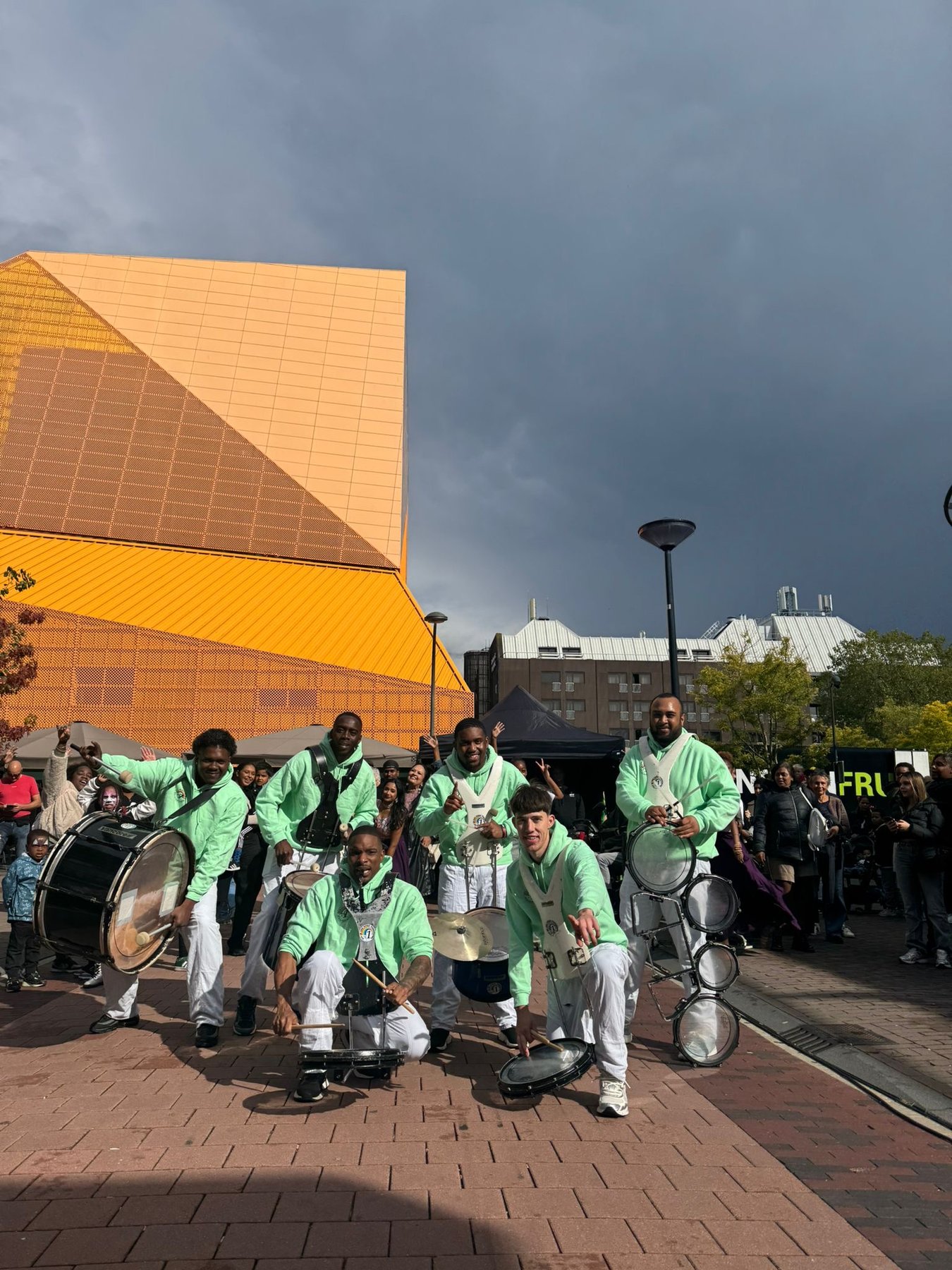 All In 1 drumband speelt buiten voor een opvallend geel-oranje gebouw